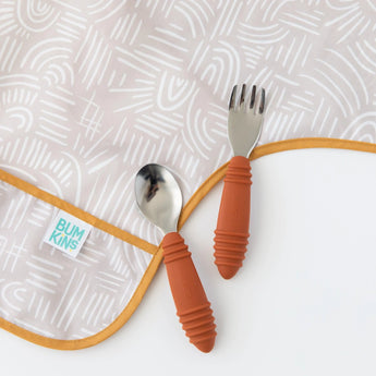 A spoon and fork with orange rubber handles sit on the patterned Sleeved Bib: Wander by Bumkins. It has waterproof fabric, a yellow trim, and a label. Ideal for toddlers, it seamlessly complements the Desert Boho style.