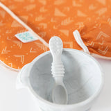 White baby spoon in a marble bowl with Bumkins Sleeved Bib: Grounded from the Desert Boho collection in the background.