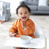 Smiling toddler in Bumkins Sleeved Bib: Grounded sits in a highchair with a spoon and bowl, ideal for baby-led weaning.