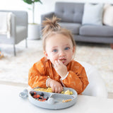 A toddler in a cozy room enjoys baby-led weaning from a divided plate, wearing a Bumkins Sleeved Bib: Grounded at a white table.