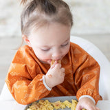 A child using a Bumkins Sleeved Bib: Grounded from the Desert Boho collection, enjoys baby-led weaning by eating with their hands.