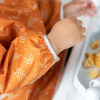 Child in a Bumkins Sleeved Bib: Grounded, seated at a table, enjoying a baby-led weaning adventure.