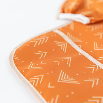 Close-up of the Bumkins Sleeved Bib: Grounded in orange fabric with white geometric patterns and cream trim from Desert Boho collection.