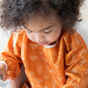 A curly-haired child in a Bumkins Sleeved Bib: Grounded sits ready for baby-led weaning.