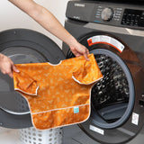 Hands hold the Bumkins Sleeved Bib: Grounded, featuring white patterns, in front of an open washing machine.