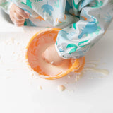 A small hand plays with food in an orange bowl, wearing Bumkins Sleeved Bib: Dinosaurs—perfect for baby-led weaning.