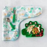 Bumkins Sleeved Bib: Dinosaurs with a green dino plate and utensils on a light surface. Ideal for baby-led weaning!.