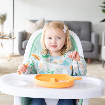 A light-haired toddler in a high chair, wearing a Bumkins Sleeved Bib: Dinosaurs, holds a fork with a yellow divided plate in front.