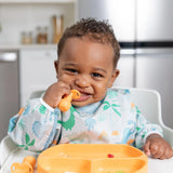 A baby in a Bumkins Sleeved Bib: Dinosaurs sits happily in a high chair with a spoon and orange plate.