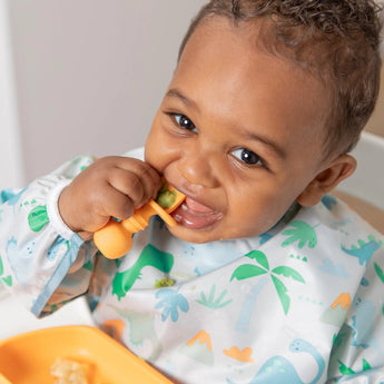 A smiling baby joyfully eats with an orange spoon and plate, wearing a Bumkins Sleeved Bib: Dinosaurs at the table.