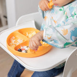 A toddler in a high chair uses an orange divided plate wearing a Bumkins Sleeved Bib: Dinosaurs while eating with a spoon.