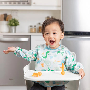 A joyful toddler in a high chair, wearing a Bumkins Sleeved Bib: Dinosaurs, raises arms excitedly during baby-led weaning in the kitchen.