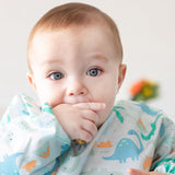 A baby with big eyes wears a Bumkins Sleeved Bib: Dinosaurs, sucking their thumb thoughtfully as blurred flowers frame the scene.