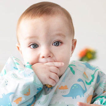 A baby with big eyes wears a Bumkins Sleeved Bib: Dinosaurs, sucking their thumb thoughtfully as blurred flowers frame the scene.