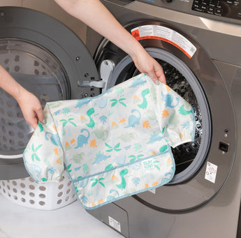 Someone puts a Bumkins Sleeved Bib: Dinosaurs, waterproof and colorful, into a front-loading washing machine.