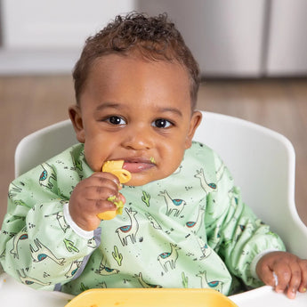 A baby in a high chair, wearing a Bumkins Sleeved Bib: Llamas, holds a piece of pasta. In the kitchen with its wooden floor and blurred background, the baby looks directly at the camera, radiating charming curiosity.