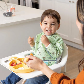 A toddler in a high chair, wearing a Bumkins Sleeved Bib: Llamas, smiles while eating from a yellow plate as an adult feeds him banana.