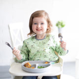 A toddler in a Bumkins Sleeved Bib: Llamas, happily holding a spoon and fork, makes clean-up easy.