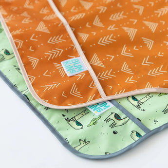 Two baby mats, one orange with white arrows and one green with dinosaurs and cacti, by Bumkins feature waterproof, easy-clean fabric.