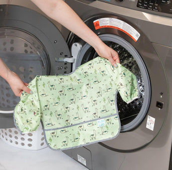 Someone loads a Bumkins Sleeved Bib: Llamas, made of waterproof fabric, into a front-loading washing machine for easy cleaning.