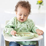 Child in a Bumkins Sleeved Bib: Llamas, made from waterproof fabric, sits in a high chair reaching for food on a green plate.