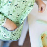 A child enjoys cereal beside the table in a Bumkins Sleeved Bib: Llamas, appreciating its easy-clean design with a pocket.