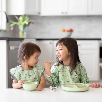 Two kids in Bumkins Sleeved Bibs: Llamas sit at a counter. One feeds the other, both smiling, enjoying easy clean-up time!.