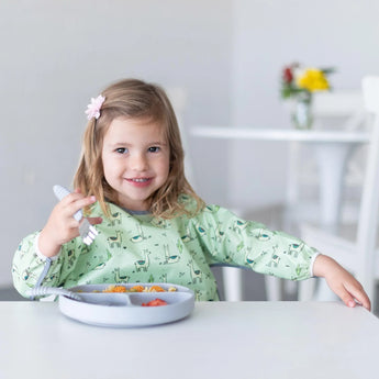 Girl in a green shirt with pink clip, wearing Bumkins Llamas sleeved bib, smiling and holding a fork at the white table.