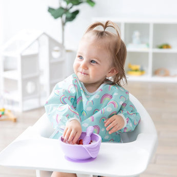 A toddler in a high chair grins while eating from a purple bowl, wearing a Bumkins Sleeved Bib: Rainbows with stain resistance.