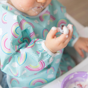 Toddler wearing a Bumkins Sleeved Bib: Rainbows, covered in yogurt and blueberries. Stain-resistant and mess-ready!.