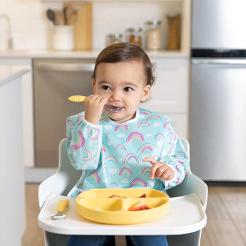 A toddler in a high chair wears a Bumkins Sleeved Bib: Rainbows, using a yellow spoon with a divided plate in front of them.