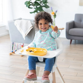 A curly-haired child in a high chair wears Bumkins Rainbows Sleeved Bib, holding food with a yellow tray in a bright room.