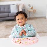 A curly-haired toddler eats fruit off a pink mat, wearing a colorful Bumkins Sleeved Bib: Rainbows, stain-resistant and joyful.