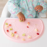 A baby in a Bumkins Sleeved Bib: Rainbows plays with food on a pink silicone tray at a highchair.