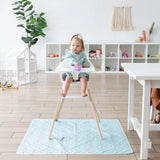 A toddler in a high chair wears a Bumkins Sleeved Bib: Rainbows, holding a cup in a bright playroom with toys.