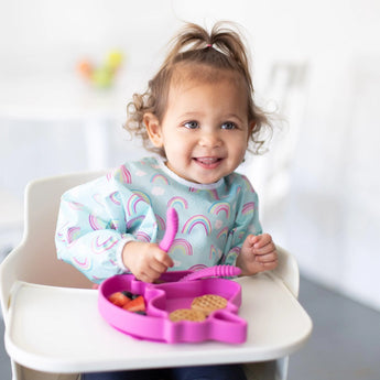 A smiling toddler in a high chair wears a Bumkins Sleeved Bib: Rainbows, holding utensils next to food on a pink plate.