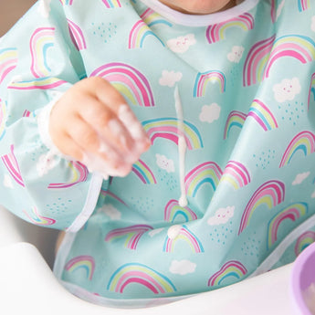 A child in a Bumkins Sleeved Bib: Rainbows reaches out, little hand smeared with food.