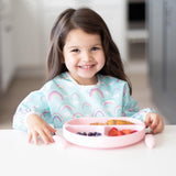 A happy girl in a Bumkins Sleeved Bib: Rainbows enjoys her meal with a pink plate.