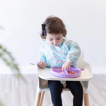 Toddler uses a Bumkins Sleeved Bib: Rainbows, eating fruit with a spoon from a purple plate in their high chair.