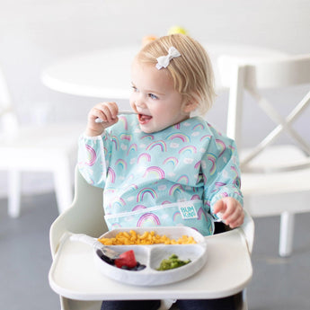 Toddler in a high chair, wearing Bumkins Sleeved Bib: Rainbows, happily enjoying pasta, strawberries, and broccoli.