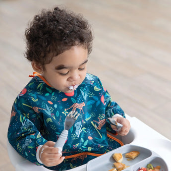 A curly-haired toddler in a Bumkins Sleeved Bib: Jungle sits in a high chair, eagerly trying baby-led weaning. Waterproof bliss!.