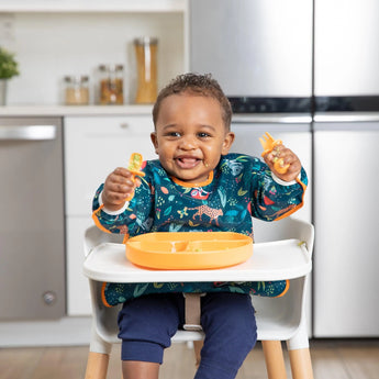 Smiling toddler in a high chair, wearing Bumkins Sleeved Bib: Jungle, holding utensils, and using a divided plate in a modern kitchen.