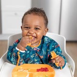 A happy baby in a high chair wears a Bumkins Sleeved Bib: Jungle while enjoying baby-led weaning with an orange plate meal.