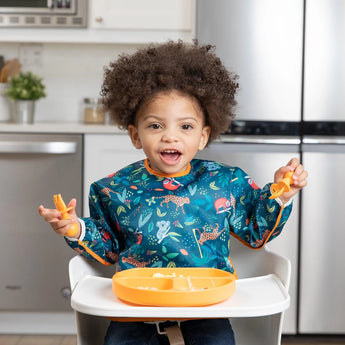 A curly-haired toddler sits in a high chair, wearing a Bumkins sleeved bib: Jungle, holding a spoon, ready for baby-led weaning.