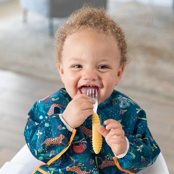 A happy toddler in a Bumkins Jungle Sleeved Bib holds a fork, embracing baby-led weaning in the high chair.