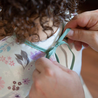 Person tying a light blue ribbon on a Bumkins Sleeved Bib: Floral. The child has curly hair.