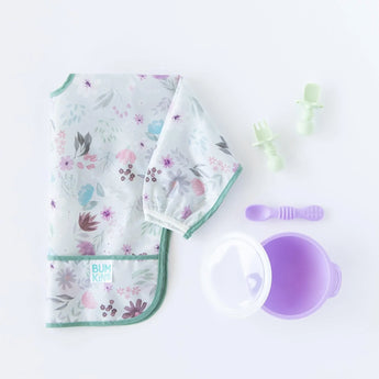 Floral sleeved bib by Bumkins, silicone spoon & fork, purple bowl with lid, and two pacifiers on white—ideal for baby-led weaning.