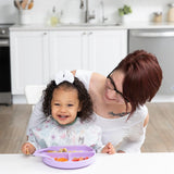 A smiling woman and toddler enjoy a meal at the table; the toddler wears a white bow and Bumkins Sleeved Bib: Floral.