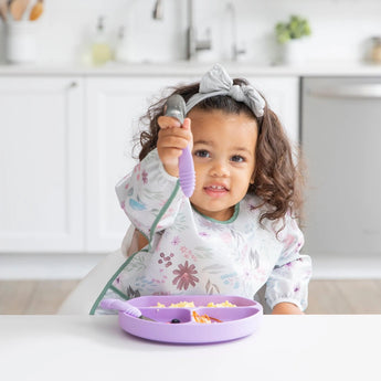 A toddler excitedly holds a spoon while wearing the Bumkins Sleeved Bib: Floral, sitting at a table with a divided purple plate.