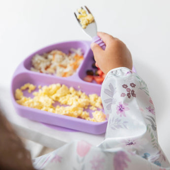 Child in a Bumkins Sleeved Bib: Floral, enjoying baby-led weaning with scrambled eggs, cheese, and veggies from a purple plate.